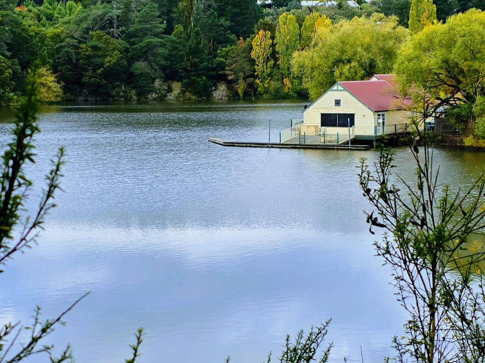 A small boathouse with a red roof on the far shore of a calm tree-lined lake
