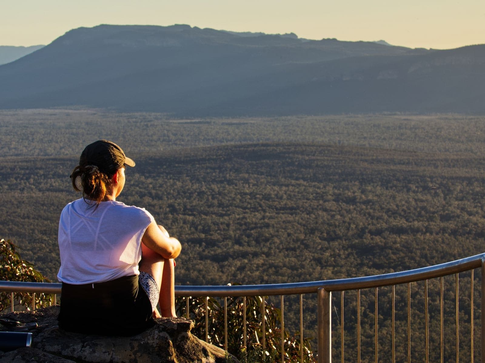 A visitor at a railed clifftop lookout gazing across forested ranges at sunset