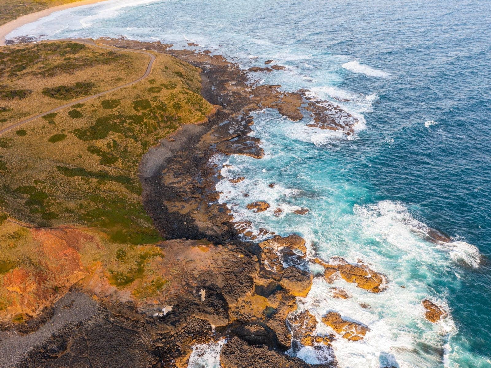 Aerial view of a rocky orange shoreline meeting clear turquoise water and white surf
