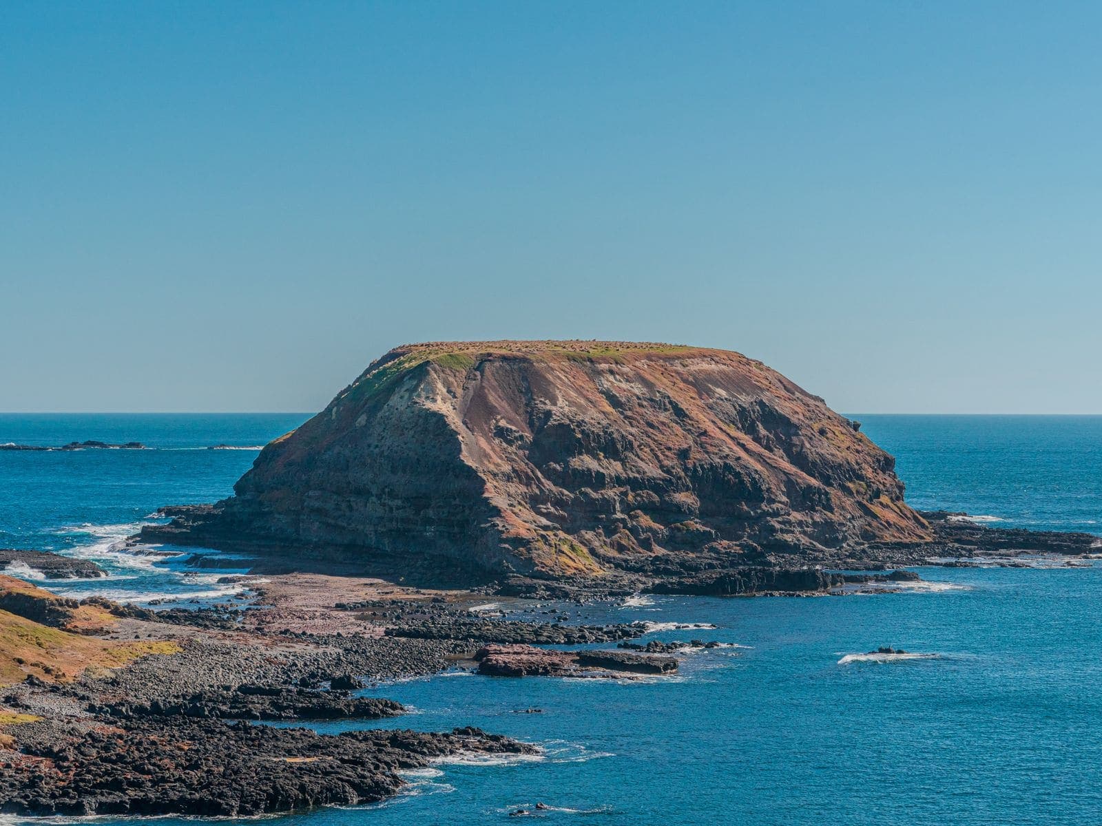 A flat-topped island of layered rock ringed by dark reef and blue ocean