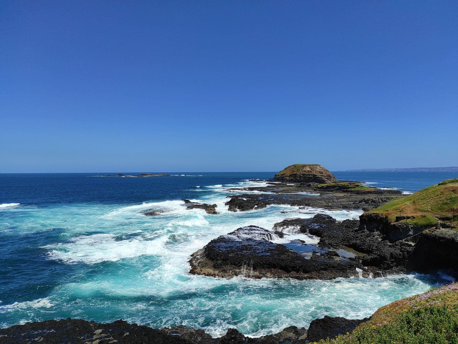 Waves breaking over a dark rocky shoreline with green headland behind