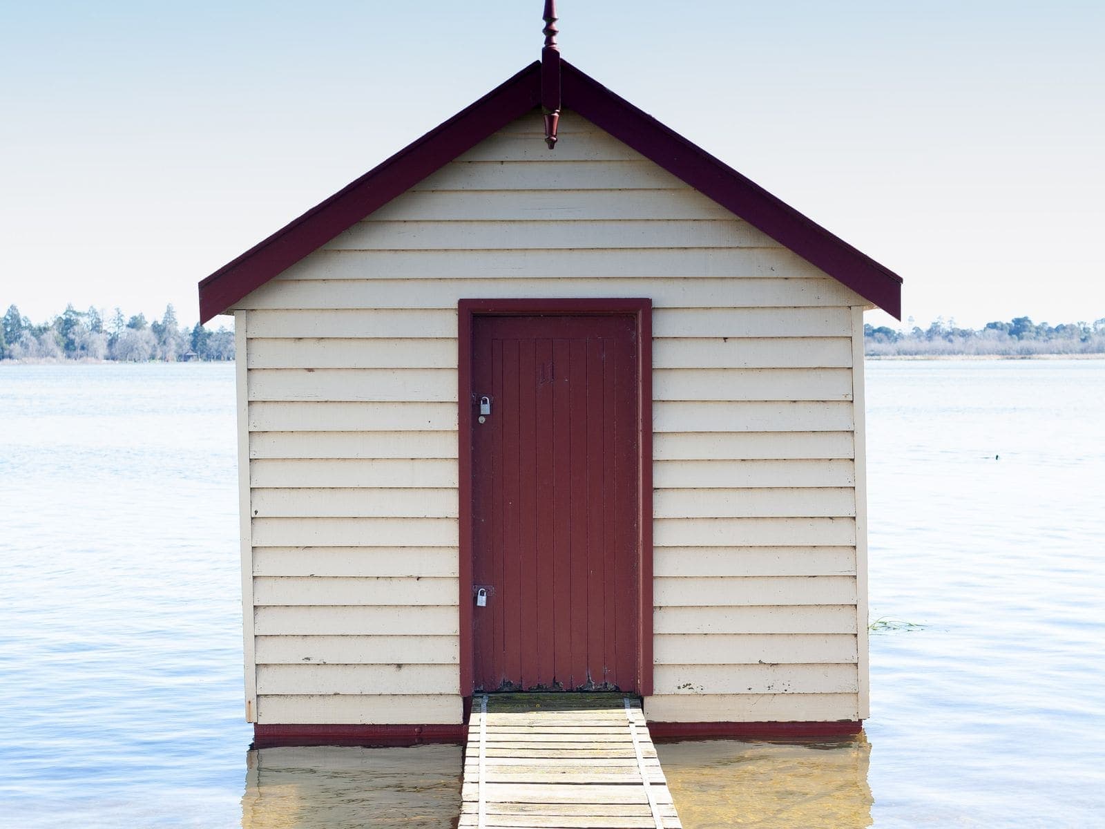 A white timber boathouse with a dark red door standing on the edge of a still lake