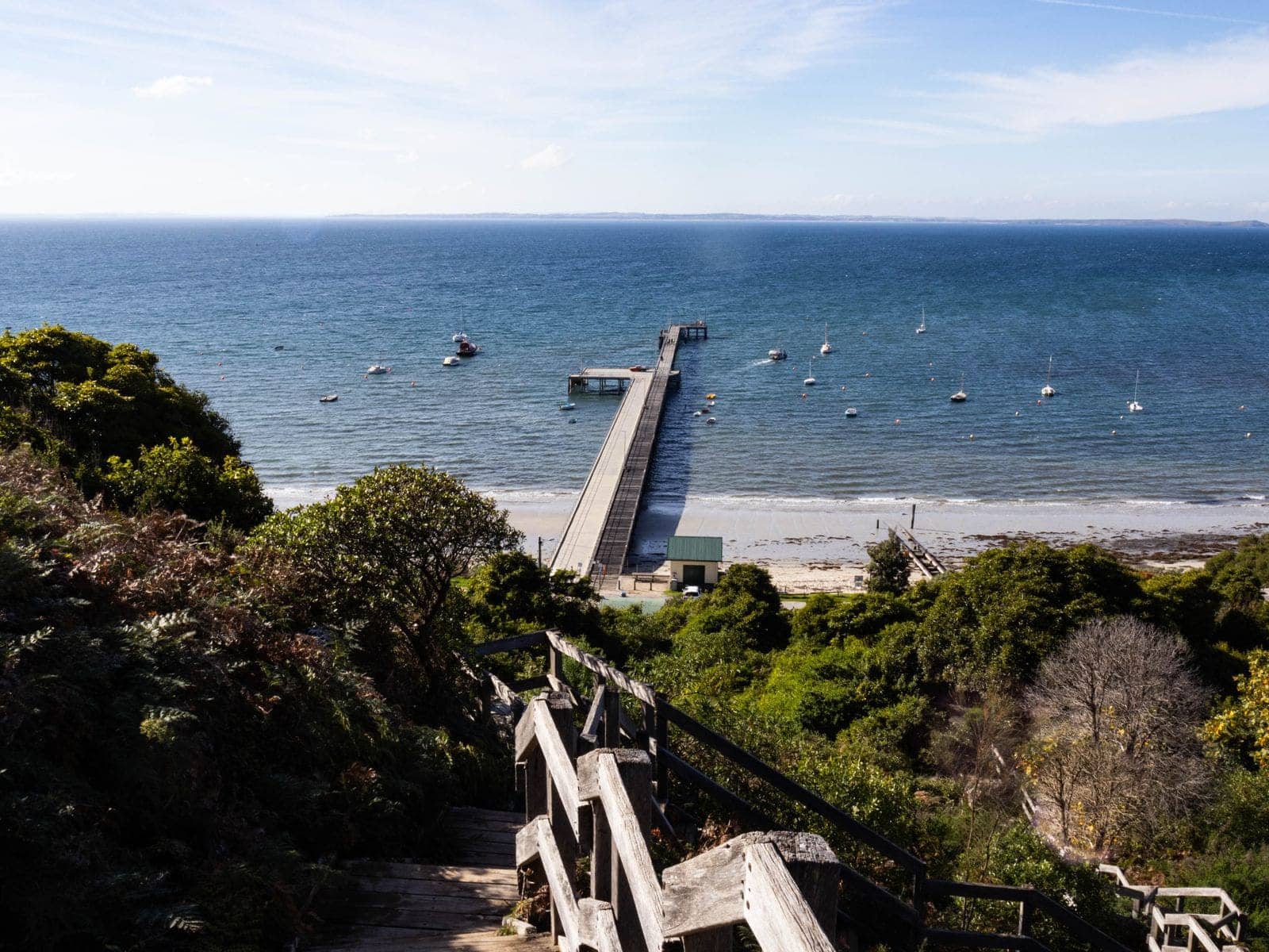 A long timber pier reaching into calm bay water with moored boats, seen from a wooded slope