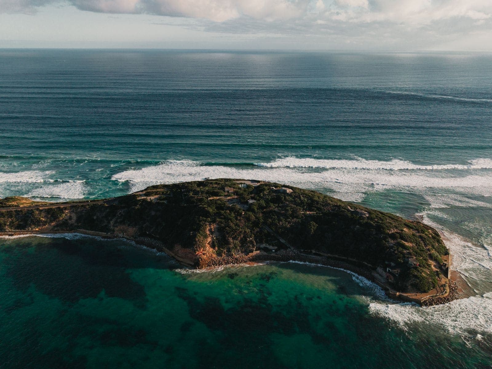 Aerial view of a green headland wrapped by turquoise shallows and breaking surf